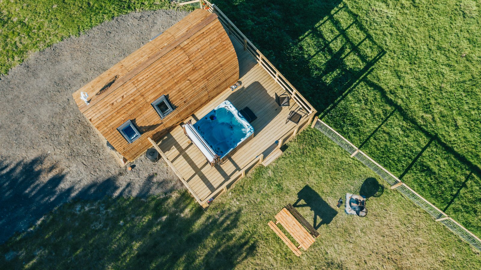 Aerial view of a small wooden cabin with a deck, featuring a hot tub and outdoor seating, nestled within a fenced grassy area in Knighton. A wooden picnic table and a fire pit are visible on the green lawn next to the cabin, perfect for those looking to experience Wigwam Holidays.