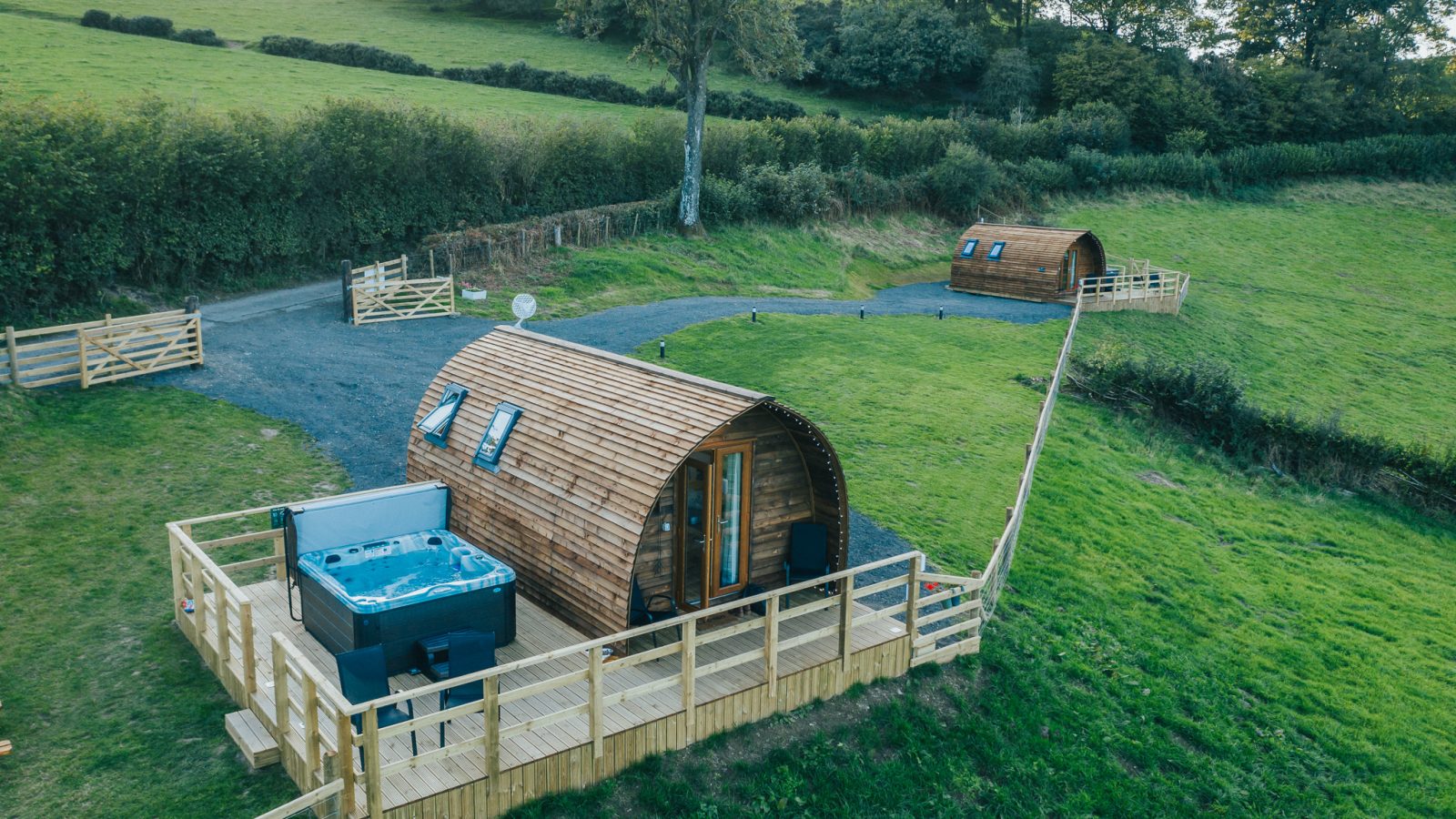 An aerial view of two wooden glamping pods situated on a lush green field at Wigwam Holidays Knighton. Each pod has a fenced outdoor deck area, with the one in the foreground featuring a hot tub. Trees and hedges border the field, enhancing the serene, rural setting.
