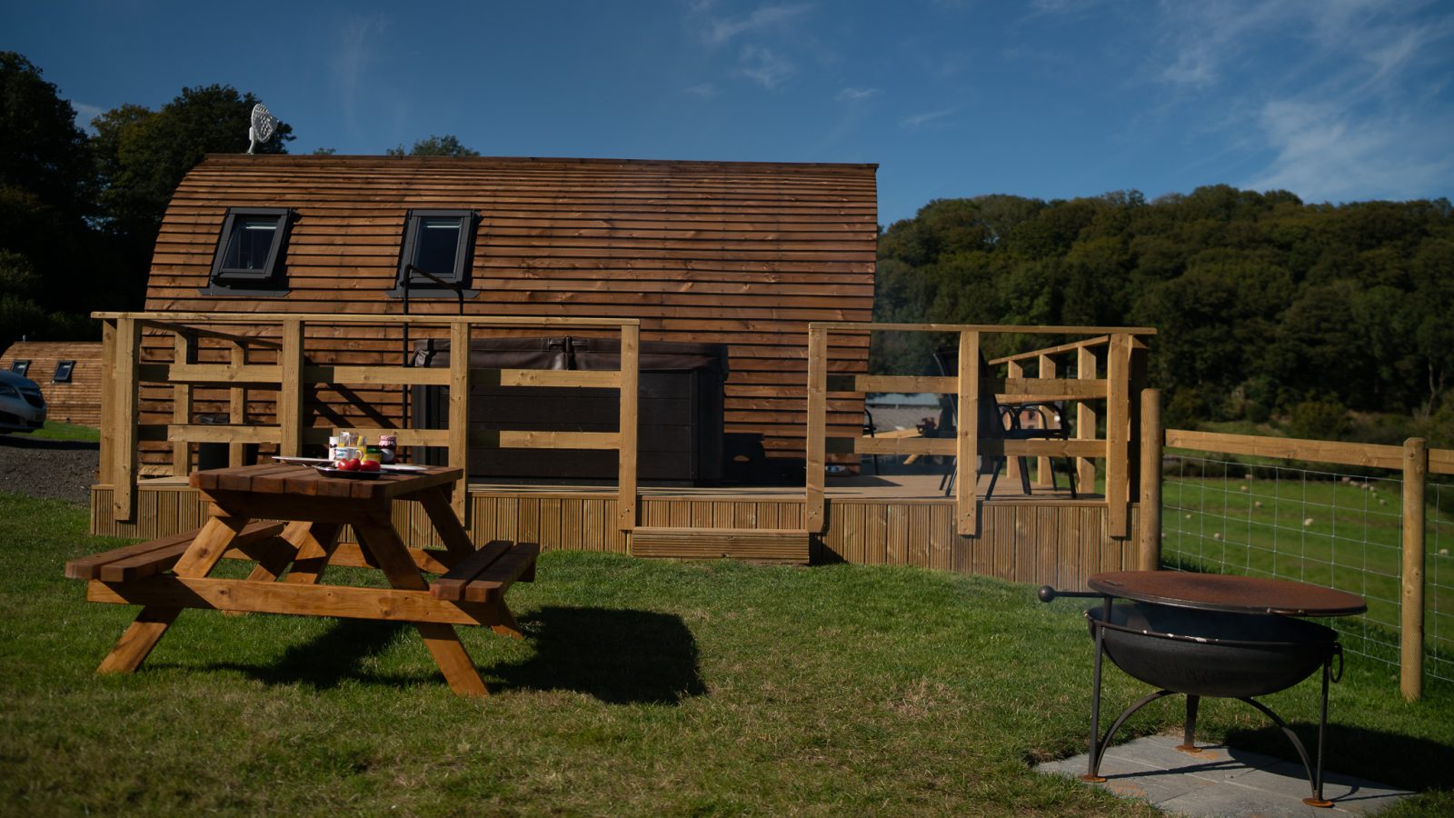 A wooden tiny house with several small windows sits on a green lawn under a blue sky. A picnic table with condiments and a fire pit are in the foreground. A wooden deck surrounds the house, and trees are visible in the background, evoking that serene Knighton ambiance perfect for Wigwam Holidays.