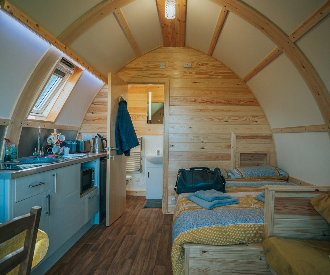 A cozy, wooden cabin interior reminiscent of a Knighton retreat features a small kitchen with a sink, stove, and microwave on the left, a dining table with chairs, and a bed with a yellow blanket on the right. The back of the cabin has a door that leads to a bathroom. The arched ceiling adds to the charm.