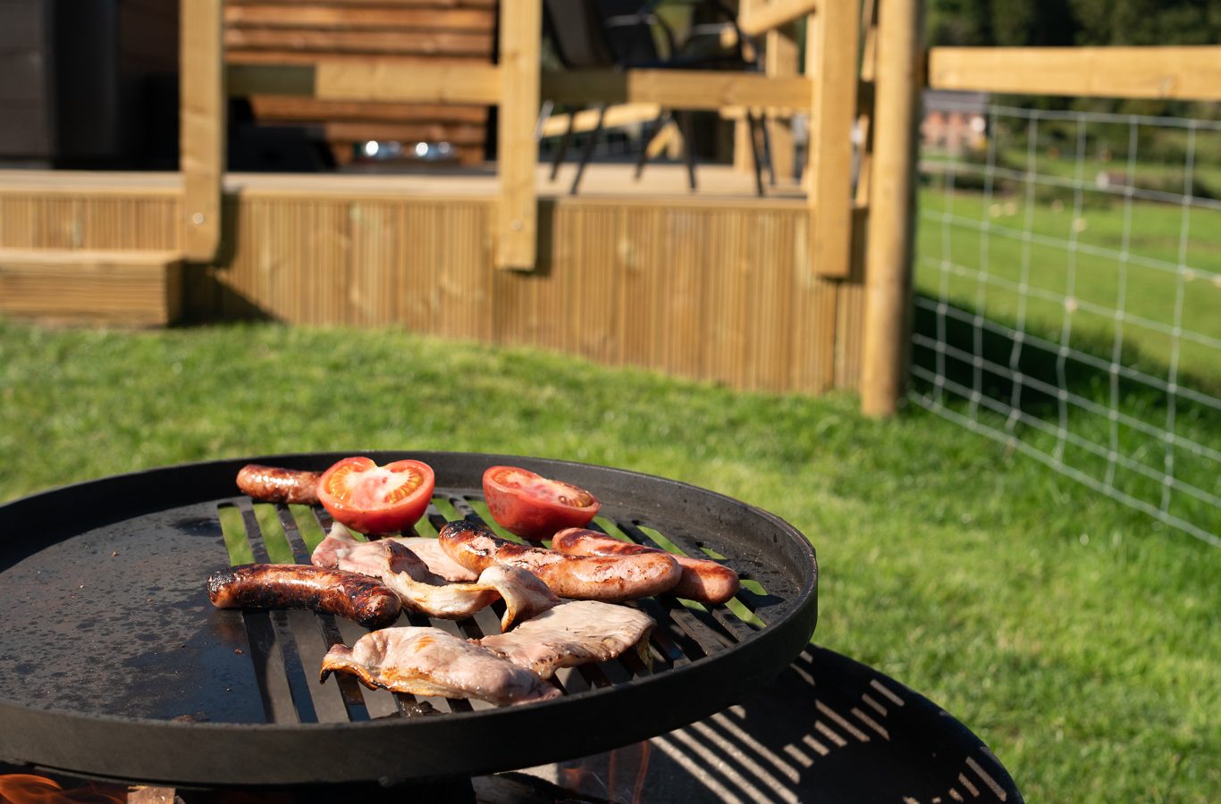 A wood-fired BBQ grill under a sunny sky with a wooden cabin and deck in the background at Wigwam Holidays Knighton. Various meats and tomatoes are grilling. The green lawn and fenced area extend towards a backdrop of trees, suggesting an outdoor cooking activity in a serene rural setting.