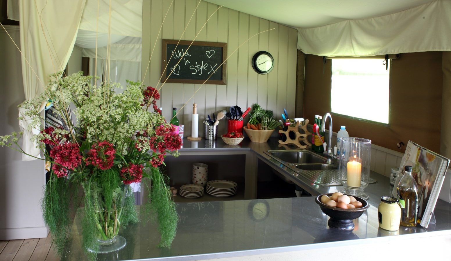 A cozy kitchen with a rustic feel featuring a flower arrangement from Drove Orchards with red and white blossoms on a shiny countertop. The space includes a sink, various kitchen utensils, and a chalkboard with the message 