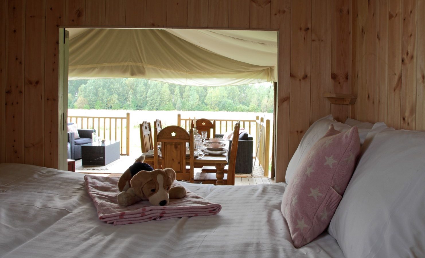 A cozy wooden cabin bedroom with a view of an outdoor deck. On the neatly made bed lies a pink pillow with star patterns and a folded blanket with a plush dog toy on top. Outside, there's a dining table and chairs, offering wild luxury as you gaze beyond at the scenic Drove Orchards forest.