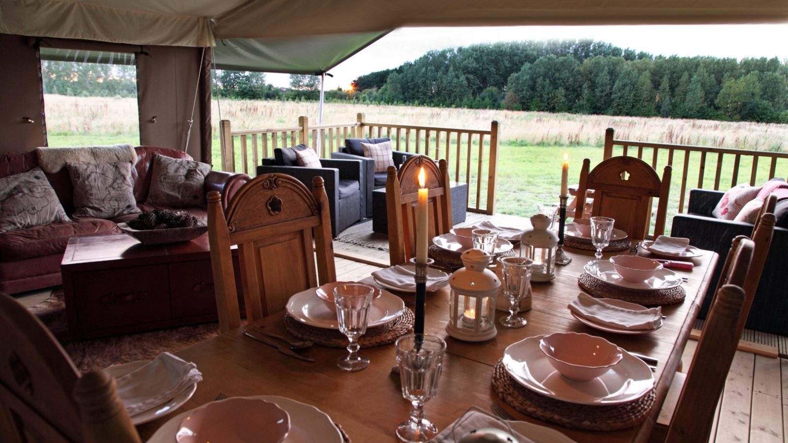 An elegant table set for a meal sits inside a spacious, furnished tent with an outdoor deck. The table has plates, glasses, napkins, and candles. The luxury seating area includes comfortable sofas, overlooking the grassy field of Drove Orchards and a line of trees in the background.