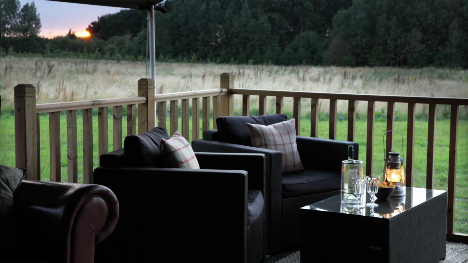 A cozy outdoor seating area on a wooden deck with two cushioned armchairs and a glass table featuring a bottle, glasses, and a lantern. The deck at Wild Luxury Drove Orchards overlooks a grassy field with trees in the background and the sun setting on the horizon.