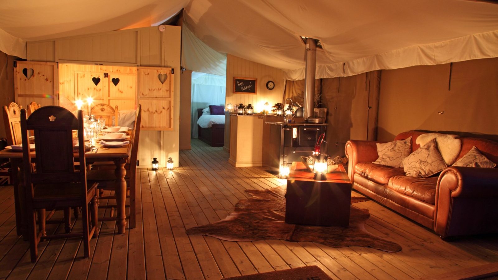 A cozy glamping tent at Drove Orchards, featuring a wooden floor and a dining area with wooden chairs and a candlelit table on the left. On the right, enjoy a seating area with a leather couch, coffee table, and soft lighting. Experience Wild Luxury with the inviting bedroom visible in the background.