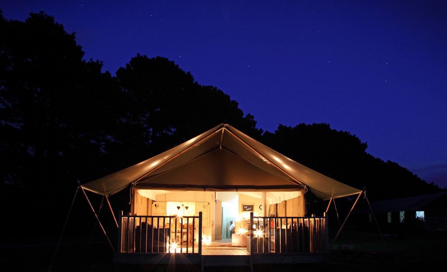 A well-lit, large canvas tent sits under a clear, starry night sky at Drove Orchards. The tent is set up on a wooden deck, surrounded by trees, and features a cozy, inviting interior with lights gently illuminating the space—a true Wild Luxury experience.