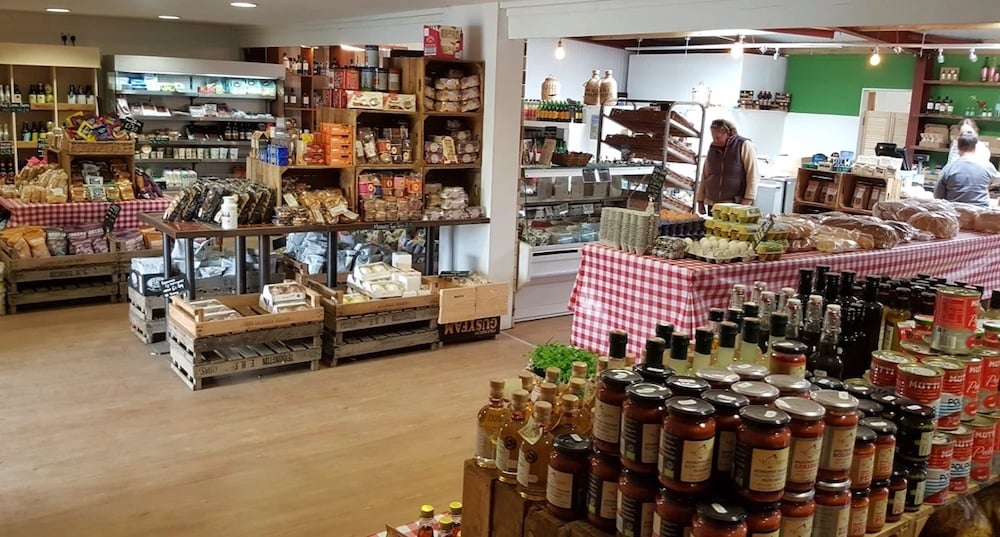 A cozy grocery store with wooden floorboards features shelves and tables stocked with various products including bottles, jars, canned goods, baked items, and fresh produce from Drove Orchards. Several customers are browsing. The atmosphere is warm, with checkered tablecloths adding a rustic touch of wild luxury.