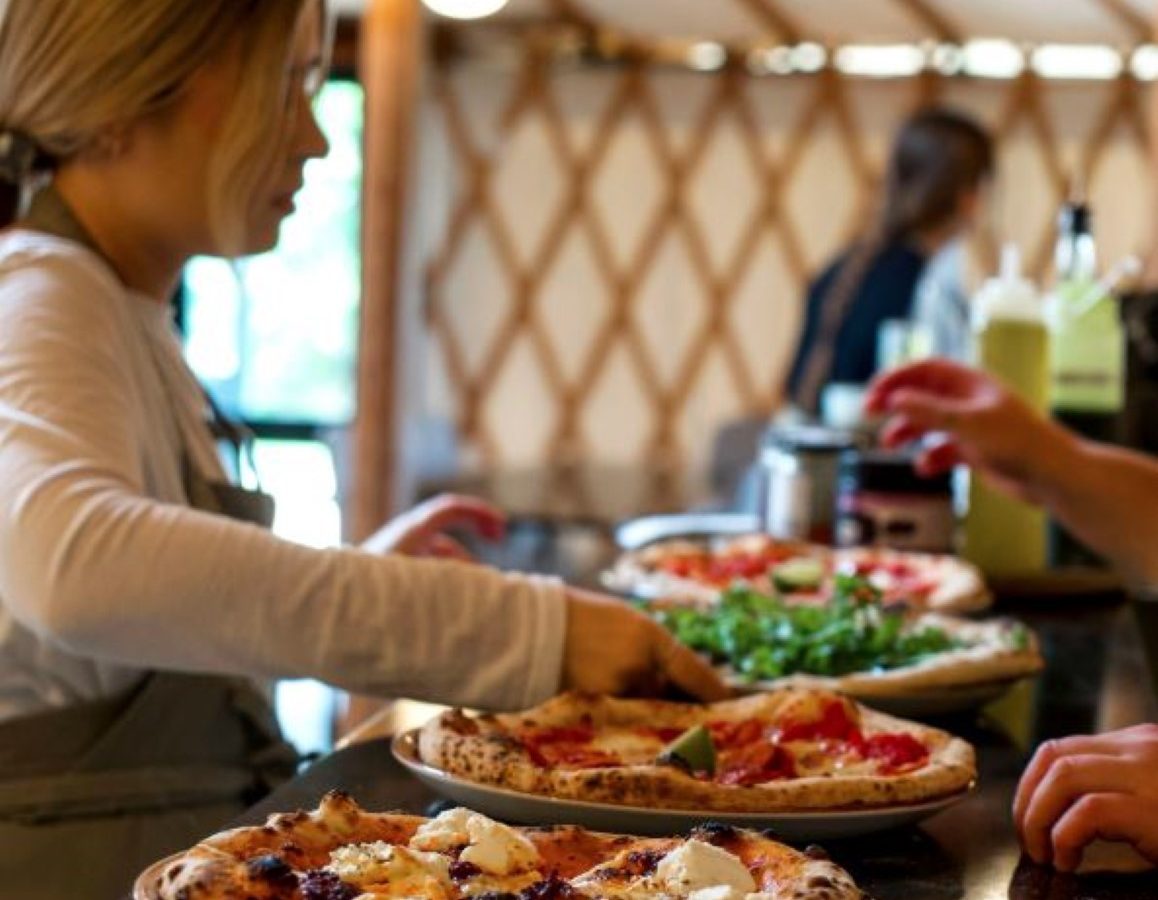 A person with light brown hair in a ponytail is serving pizza in a cozy, warmly lit restaurant at Drove Orchards. Freshly made pizzas are on the counter, and another person is working in the background. The ambiance is rustic and inviting, adding a touch of wild luxury to your dining experience.