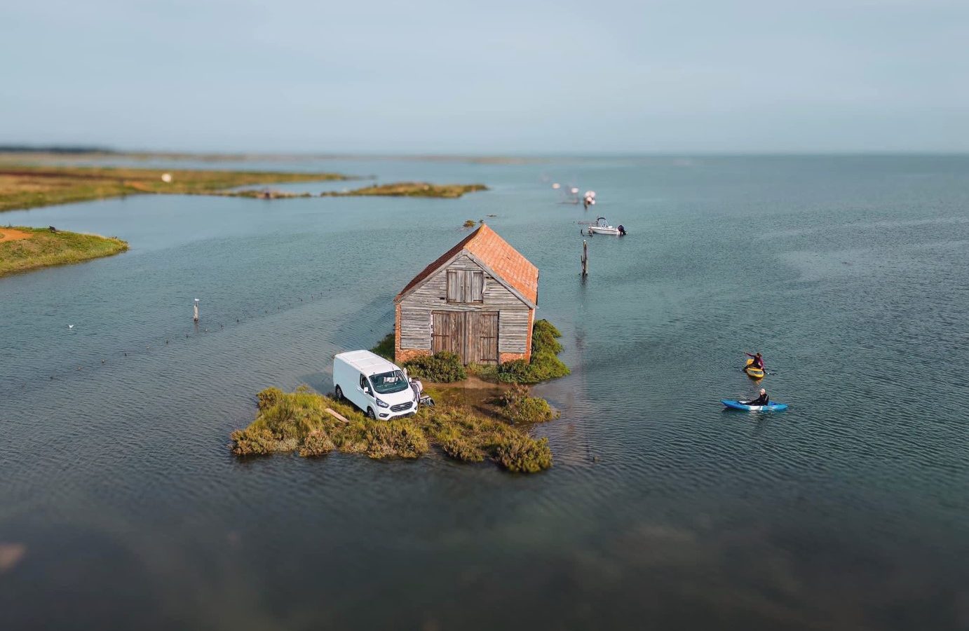 An old wooden shack with a red roof sits on a tiny, grassy islet surrounded by water. A white van is parked next to the shack, perhaps from a recent visit to Drove Orchards. Nearby, a person on a paddleboard glides across the calm waters under a clear sky, adding a touch of wild luxury to the serene setting.