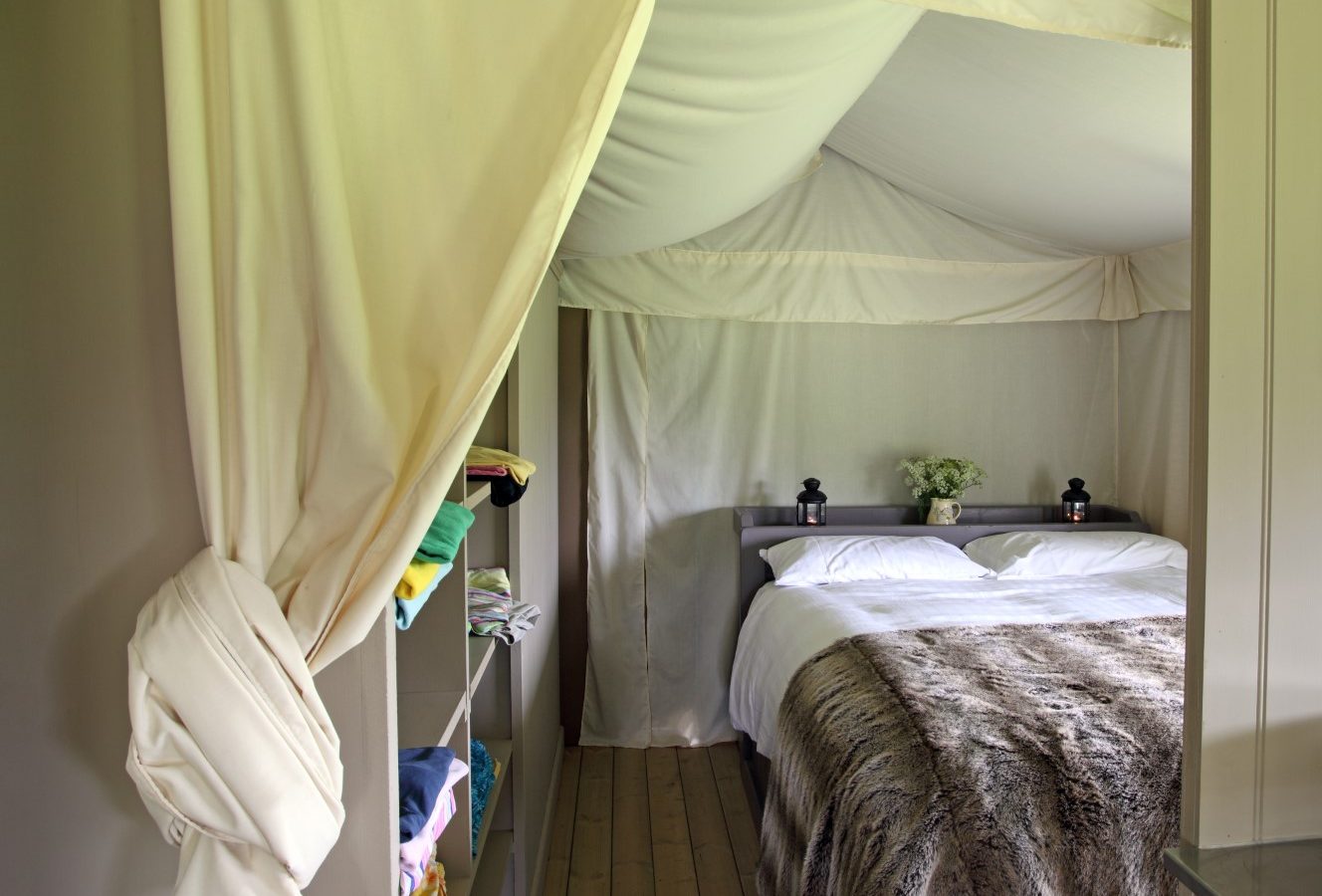 A cozy tent interior at Drove Orchards features a neatly made bed with white linens and a fur blanket. The light-colored fabric curtains and walls add to the Wild Luxury ambiance. Shelving on the left holds colorful folded towels, while two small lamps and a vase with flowers sit on the bedside table.