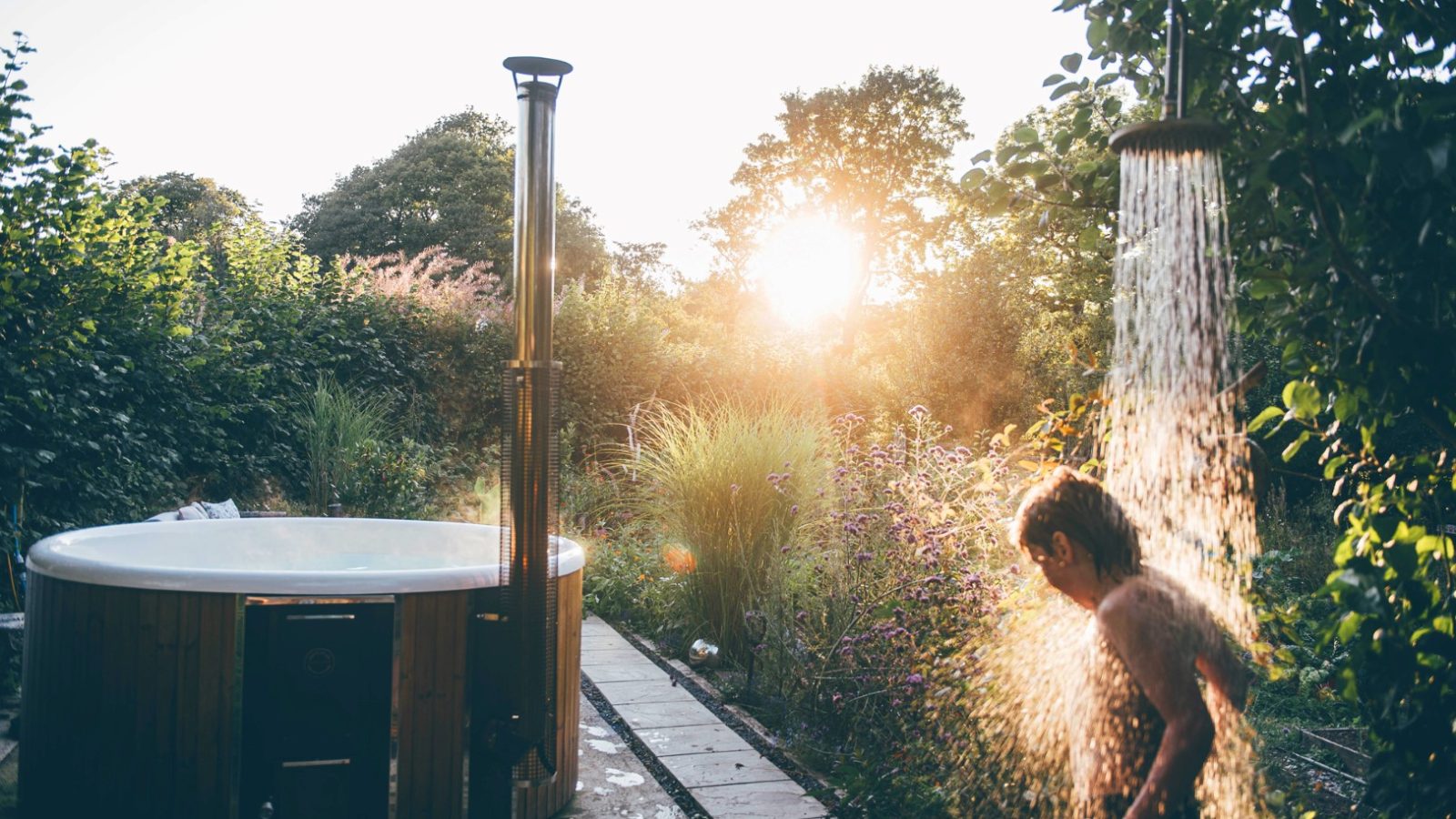 A person takes an outdoor shower beside a wooden hot tub in a lush garden, illuminated by the setting sun. Tall trees and dense greenery surround the serene Nantseren scene, creating a peaceful and natural atmosphere.