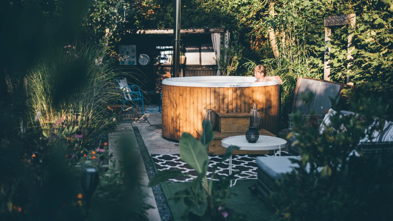 A person relaxes in a wooden hot tub surrounded by lush greenery in a Nantseren-inspired garden. The area features various plants, outdoor furniture, and a cozy atmosphere with a mix of natural and modern elements. The scene is tranquil and inviting.