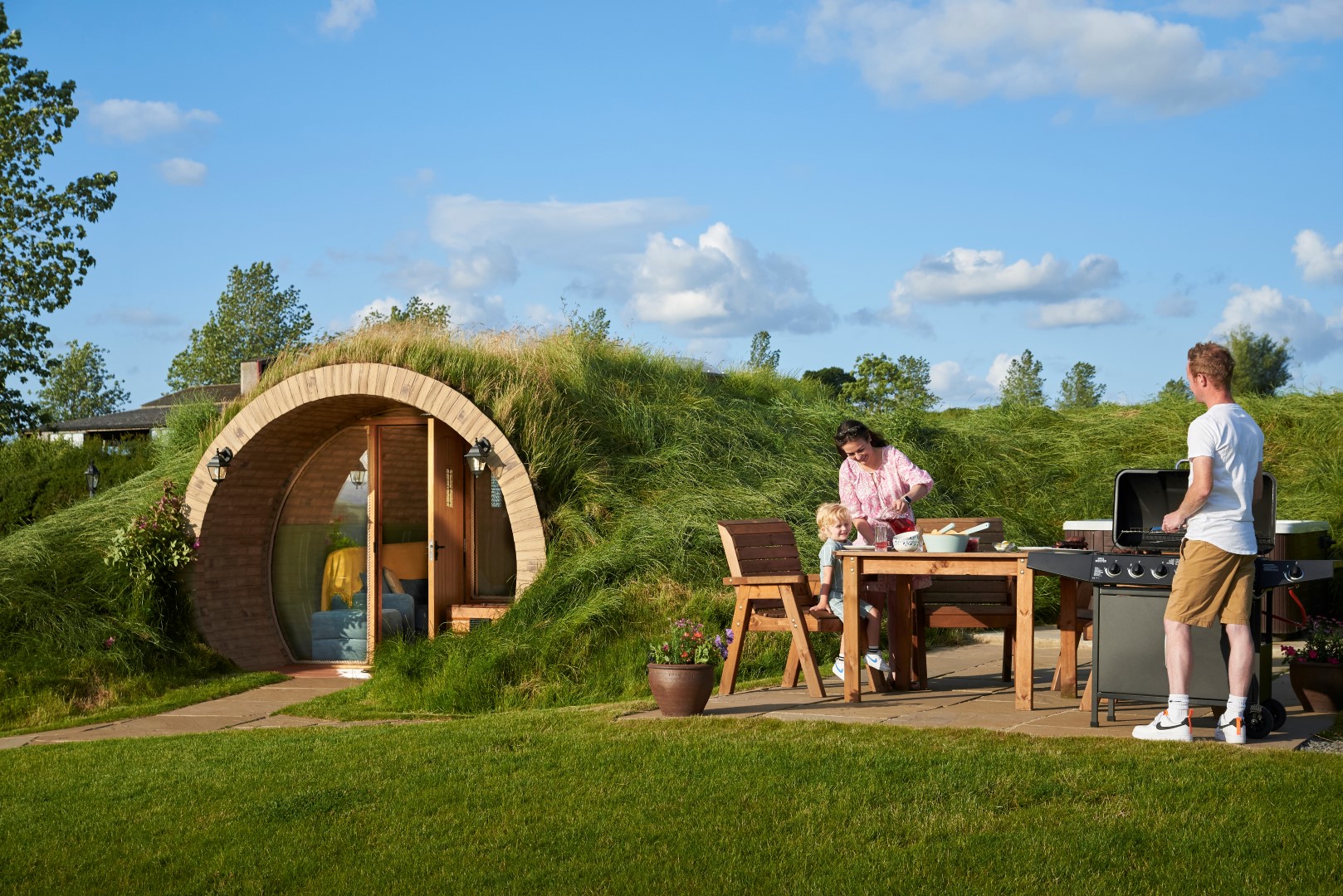 A family enjoys a sunny day outside their grass-roofed, hobbit-style house. A man grills food on a barbecue, while a woman and a child prepare a meal at a wooden table. The grassy landscape and blue sky with scattered clouds create a serene and picturesque setting.