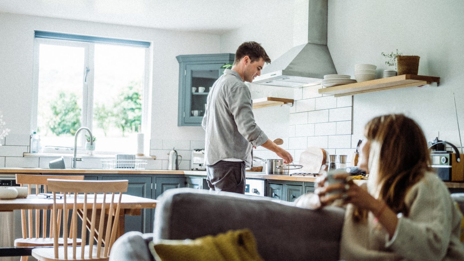 A man is in the kitchen, washing dishes at the sink. In the foreground, a woman sits on a couch holding a mug, partially turned to look at him. The kitchen is bright with natural light from a window, with modern decor featuring blue cabinets and wooden shelves reminiscent of a cozy lodge.