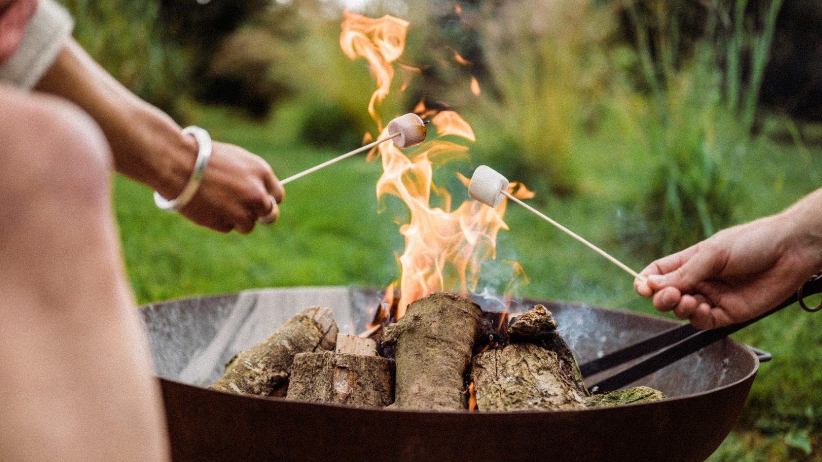 Several people roast marshmallows over a fire pit in the serene outdoor setting of Owl Lodge. The marshmallows are held on long skewers, with flames rising from the burning wood in the pit. The background features green foliage and a blurred natural landscape.