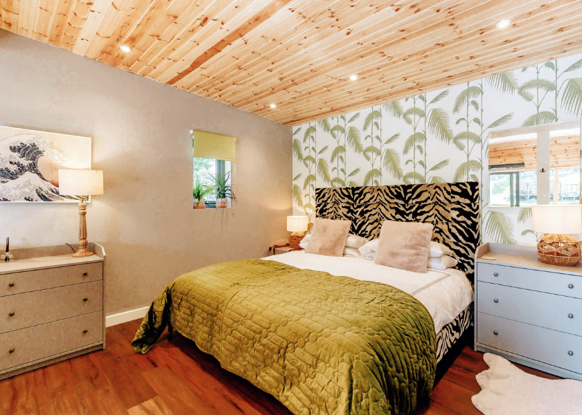 A cozy bedroom reminiscent of a Blossom Plantation, featuring a wooden ceiling and palm-leaf wallpaper. It boasts a large bed with a patterned headboard and green quilt, flanked by bedside tables lit by lamps. Natural light filters through the window with a green shade.