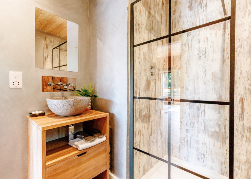 A modern bathroom with a wooden vanity featuring a stone sink, mirror, and decorative items. The space is reminiscent of a Blossom Plantation, with a large shower showcasing a rustic wood-textured wall and black-framed glass door. Natural light floods the pods of tranquility within.