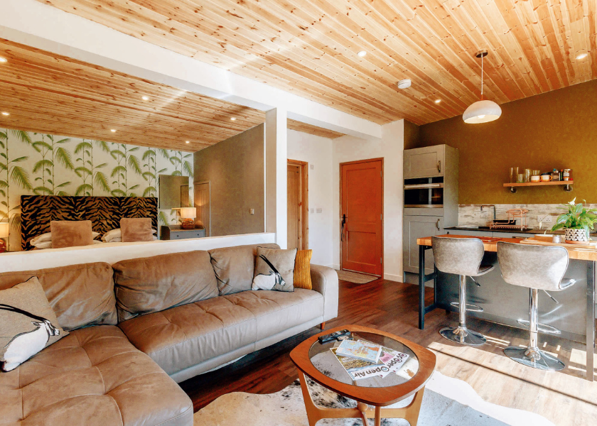 A cozy living space with a large brown sofa, a wooden coffee table, and a small kitchen area. Wooden ceilings complement the bed's patterned wallpaper, while a door leads to another area filled with warm, natural lighting—perfect for watching blossoms from nearby plantations thrive.