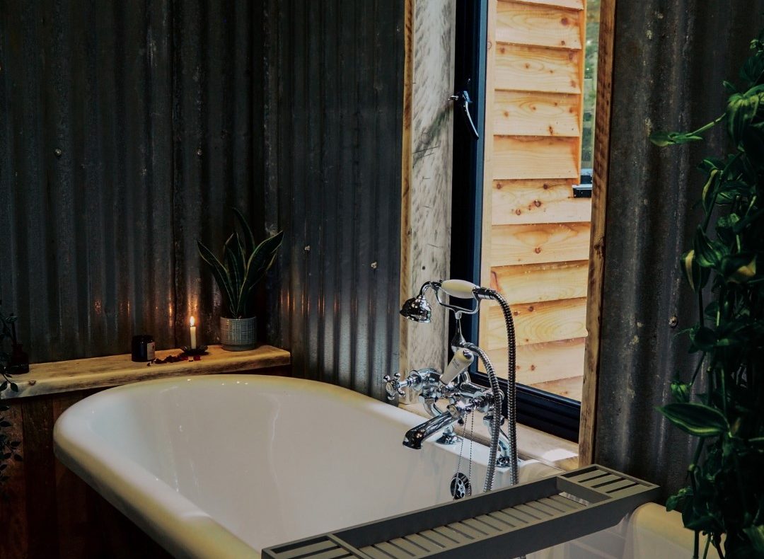 A cosy bathroom in this charming treehouse within 2 hours of London features a white freestanding bathtub against a corrugated metal wall. A wooden shelf holds a potted plant and candle beside the tub. A window with wooden panelling looks outside, with another plant visible in the foreground.