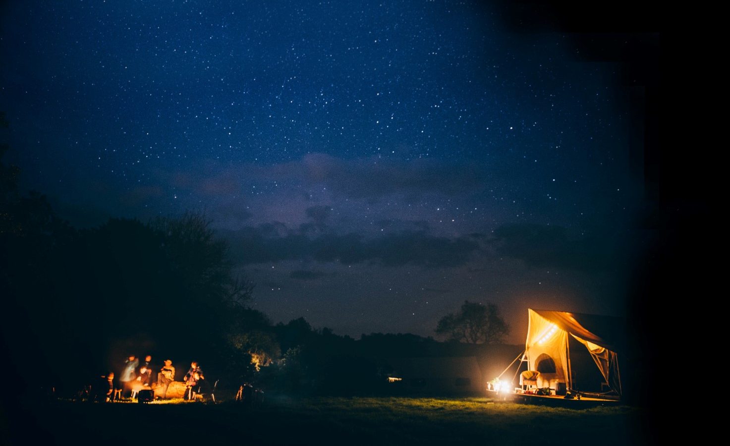 A nighttime scene shows a group of people gathered around a campfire under a Nantseren star-filled sky. In the distance, a brightly lit tent stands out against the dark surroundings. Trees and clouds are faintly visible in the background.