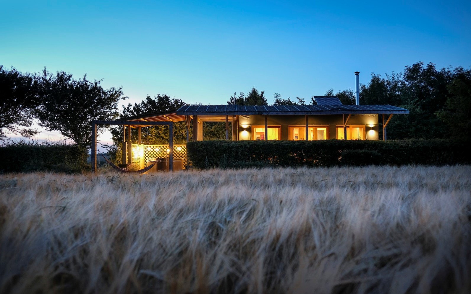 A small cabin with illuminated porch lights is surrounded by tall grass and trees at dusk, offering a serene retreat among places to stay within 2 hours of London.