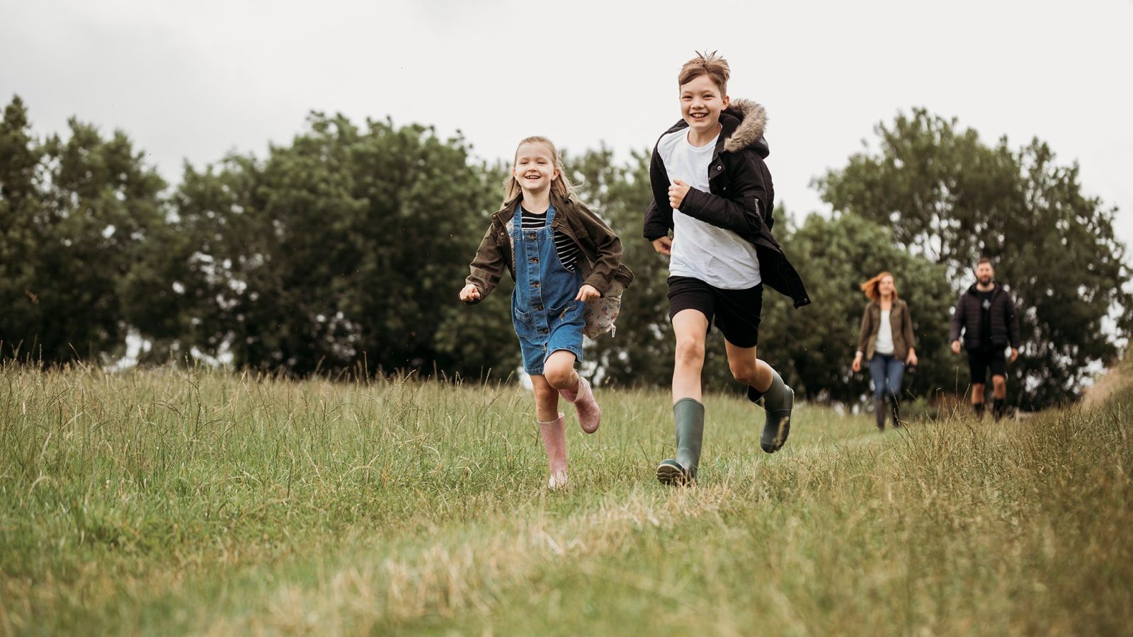 Two children, a boy and a girl, joyfully running on a grassy field. The boy is wearing a black coat and green boots, while the girl is in a blue dress and pink boots. In the background, four adults stroll near trees under a cloudy sky, with a safari tent visible in the distance.