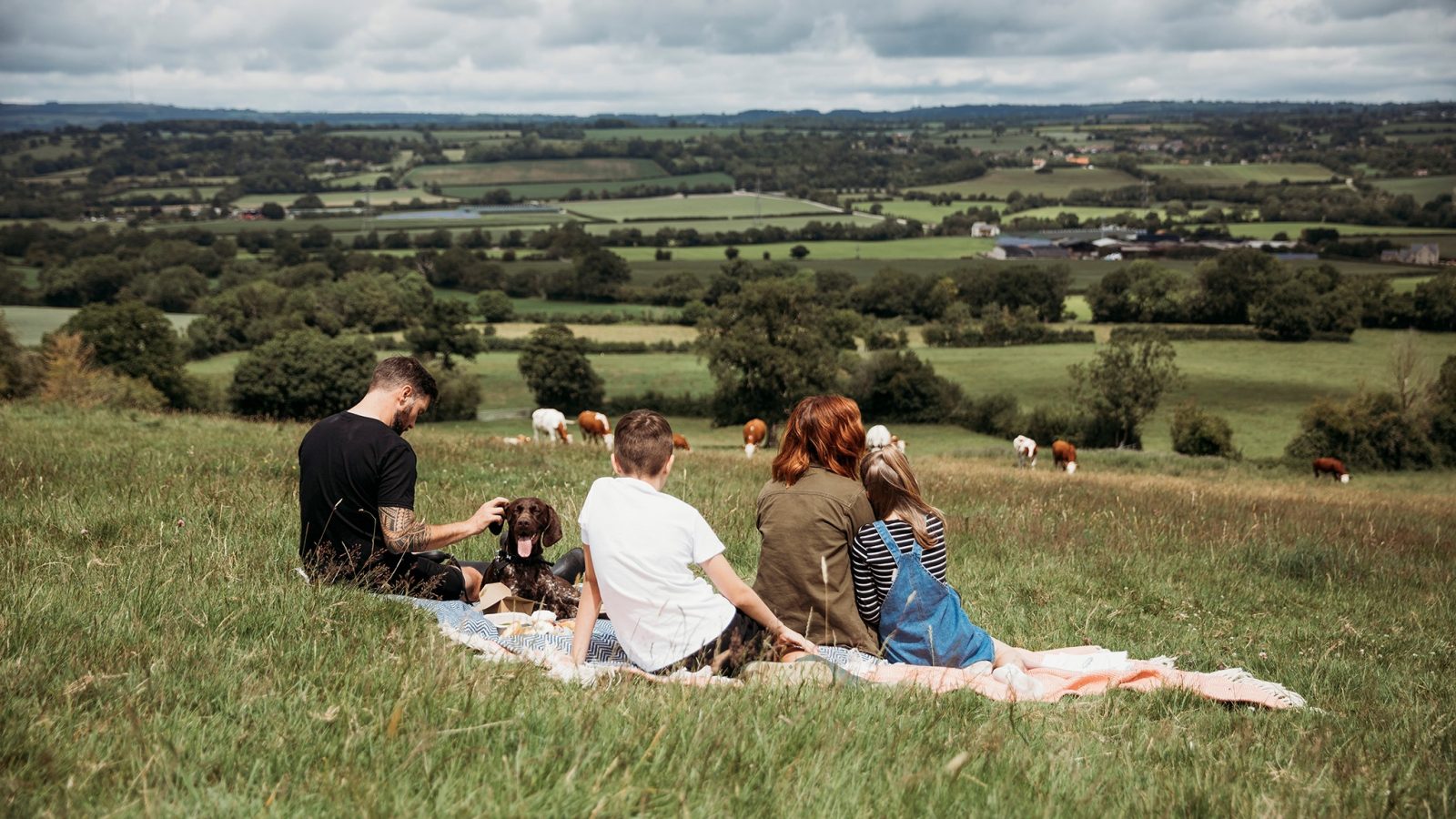 A family of four, including a man, two children, and a woman, sits on a blanket having a picnic near their safari tent on a grassy hill. They face away, looking at a scenic view of green fields and grazing cows. The sky is partly cloudy, and the landscape stretches into the distance.