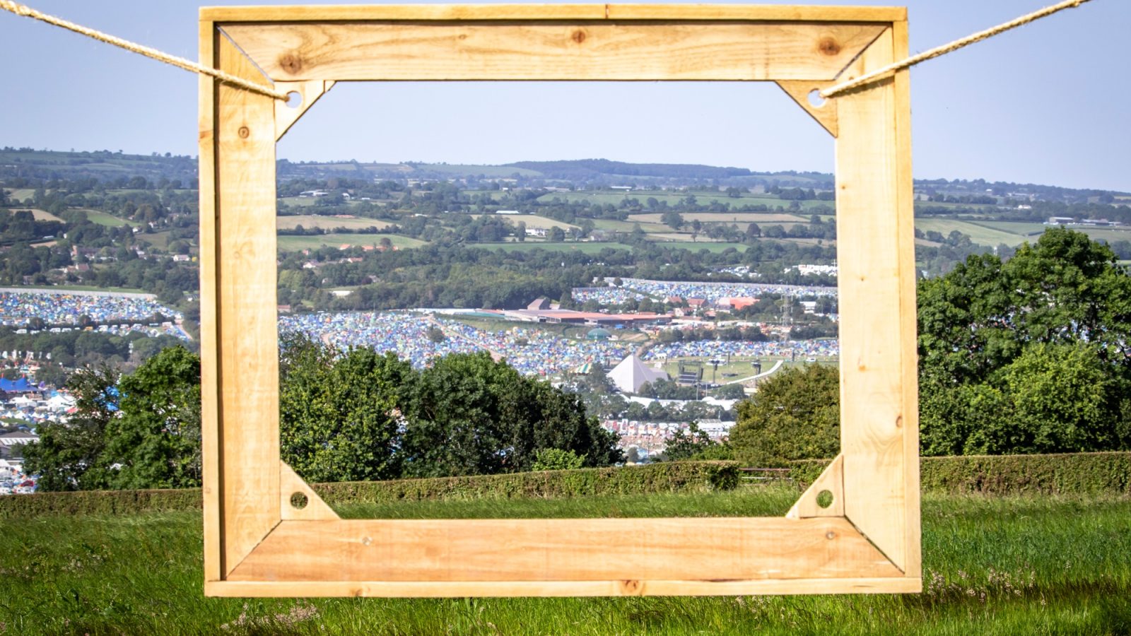 A wooden picture frame is suspended by ropes against a scenic backdrop of rolling green hills and a distant town, with a nearby safari tent adding an adventurous touch. The frame appears to highlight the landscape within it, giving the effect of a framed photograph of the picturesque view.