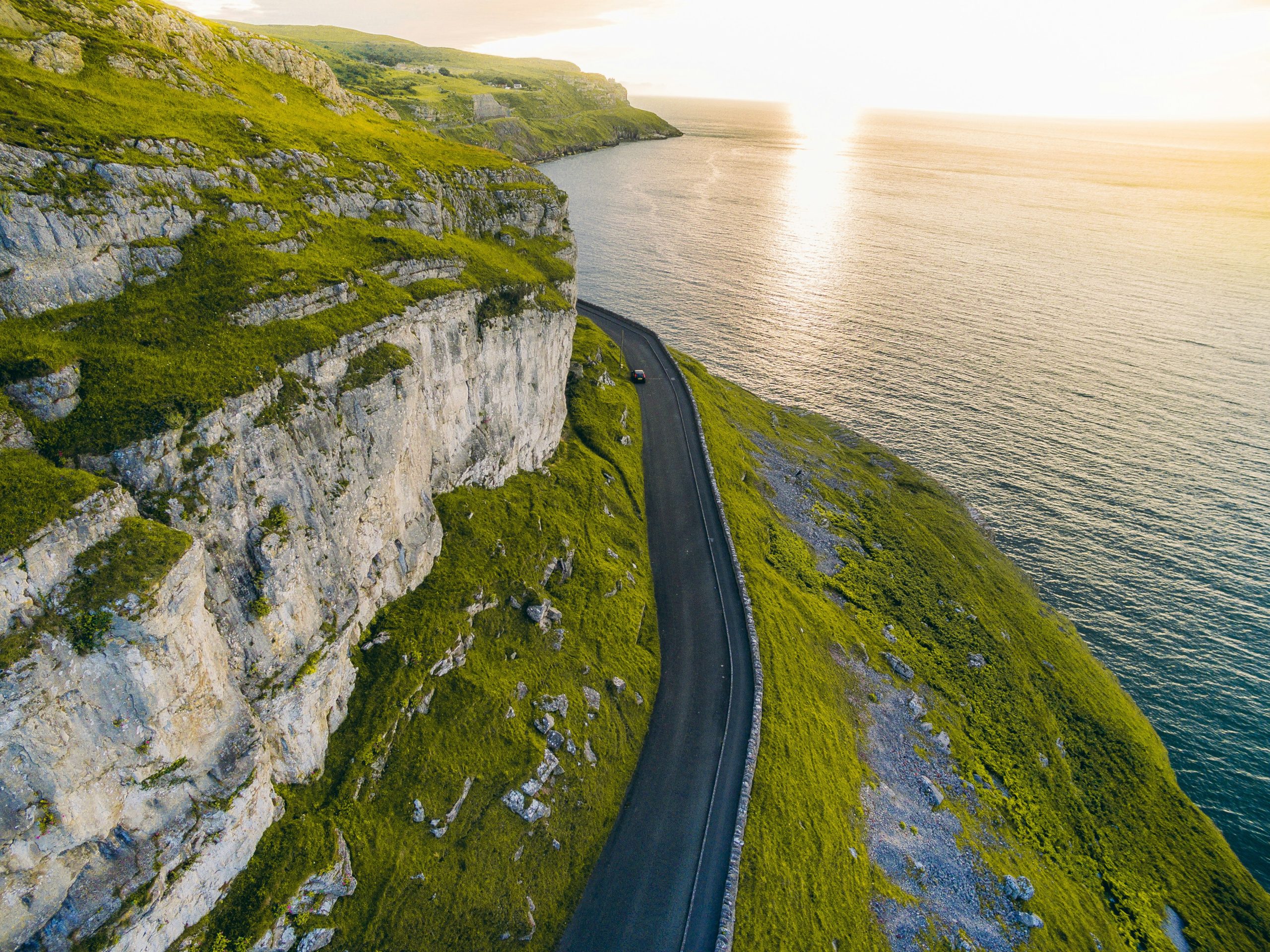 A winding road in North Wales runs along a cliffside, the setting sun casting a warm glow over the ocean's landscape.