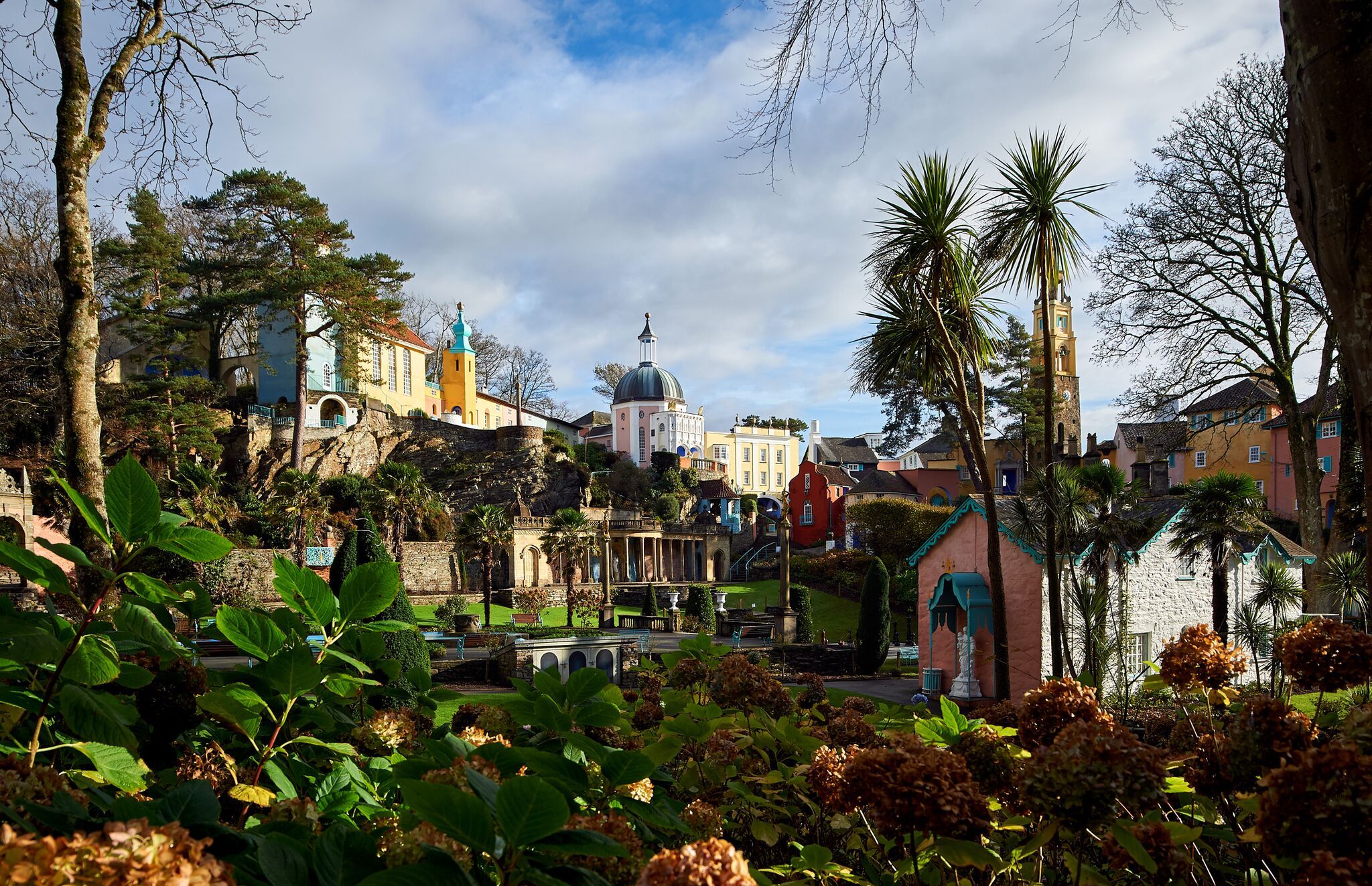 Scenic view of Portmeirion, a must-see destination when considering where to go in North Wales, with its colorful buildings, lush greenery, and clear sky framed by trees and vibrant flowers.