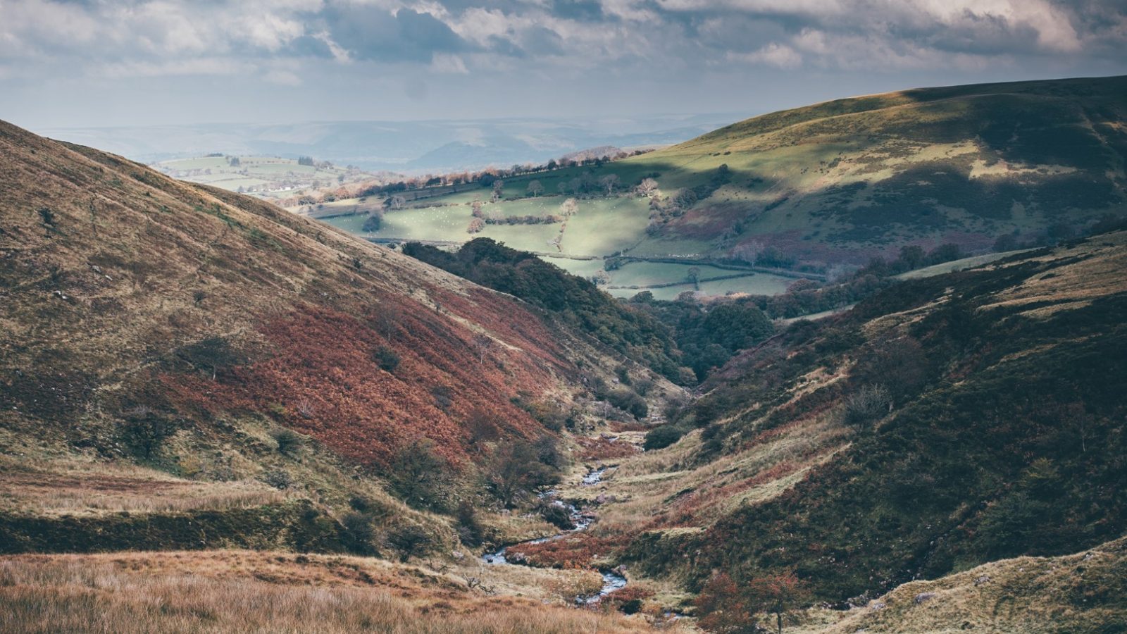 A scenic view of Nantseren Valley with a narrow stream flowing through it, surrounded by grassy hills. Some slopes are covered with reddish-brown vegetation, while others are lush green. The sky above is partly cloudy, casting shadows on the landscape.