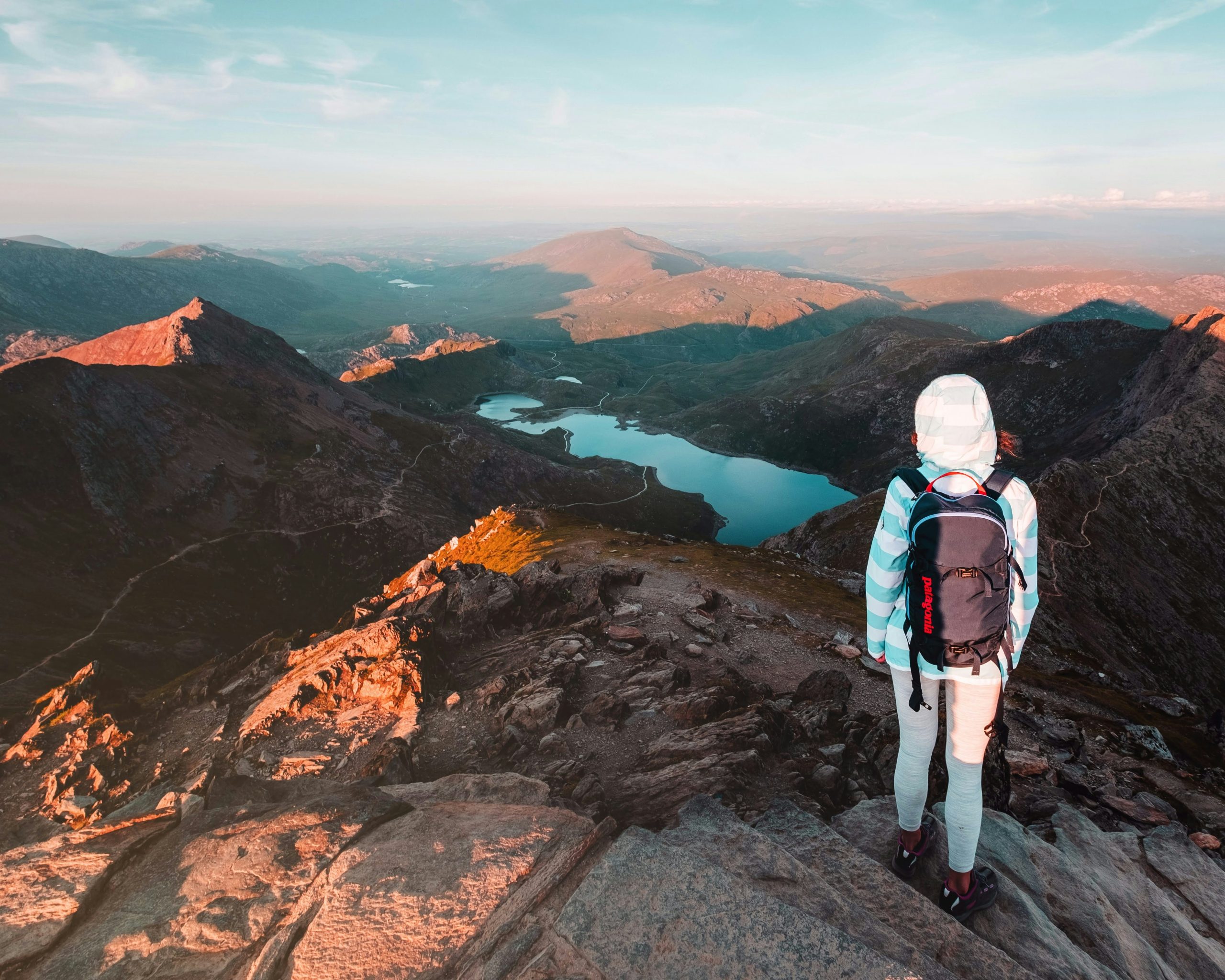 A guide to North Wales: A hiker stands atop a rocky peak, admiring the vast mountains and lake under a clear sky.