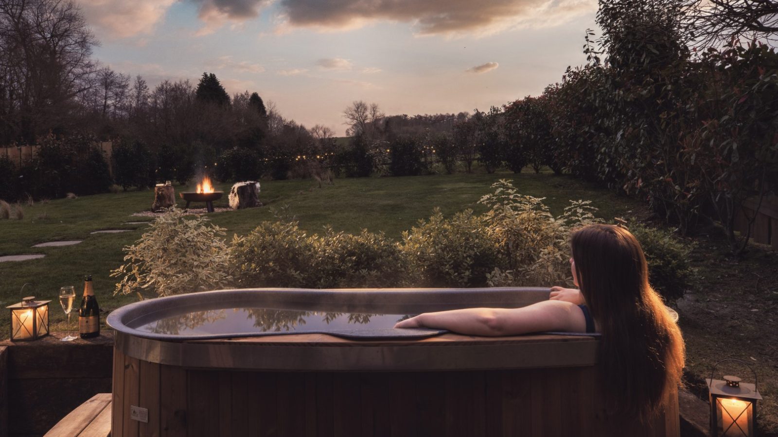 Person with long hair relaxing in an outdoor wooden hot tub at Owl Lodge, facing a garden with topiary and a fire pit under a partly cloudy sky during sunset. Lanterns and a bottle of champagne with glasses are placed next to the tub. Trees and shrubbery surround the yard.