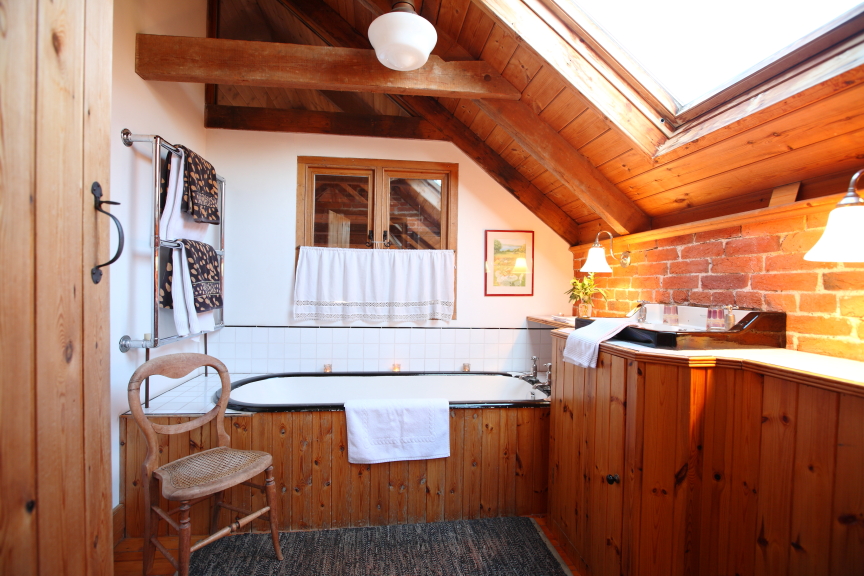 A cozy bathroom with wood-paneled walls and ceiling, a skylight, and a bathtub with white tile. The space includes a wooden chair, wall-mounted towels, hanging lights, and a small window. The decor features rustic elements reminiscent of Pennard Farm Cottages.