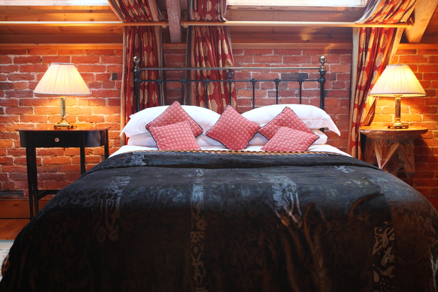 A cozy bedroom in an attic with a brick wall, skylights, and wooden beams awaits you at Pennard Farm Cottages. The bed features a wrought iron frame with a dark patterned blanket, white sheets, and four red patterned pillows. Two bedside tables with matching lamps and red curtains adorn the room.