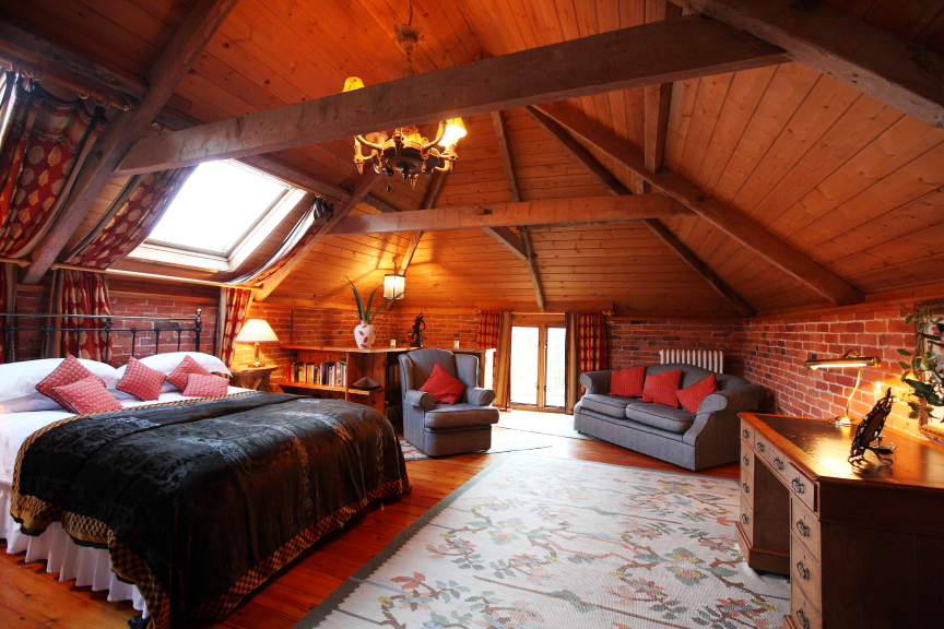 A cozy attic bedroom at Pennard Farm Cottages features a wooden ceiling and exposed beams. The room includes a bed with a black throw and red pillows, a skylight, a couch, a desk with a lamp, and a patterned rug on the wooden floor. The walls are partly brick and feature windows with curtains.