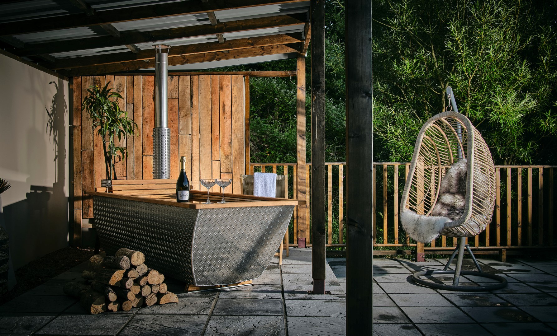 Outdoor setup featuring a lounge area with a large wood fired hottub filled with water, a bottle of wine, and two wine glasses on the edge. A cosy egg-shaped hanging chair with a fur throw is nearby. The space is surrounded by wooden fencing and lush greenery—truly one of those dreamy Devon stays with hot tubs and outdoor baths.