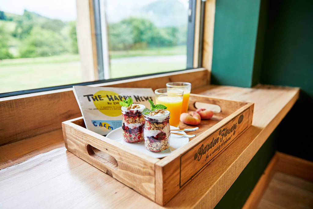 A wooden tray on a windowsill holds two jars of overnight oats with berries and mint, a glass of orange juice, a small bowl of peaches, and a magazine titled "The Happy News." The background view shows a green landscape through the window—a perfect morning to consider last-minute availability for holiday lets.