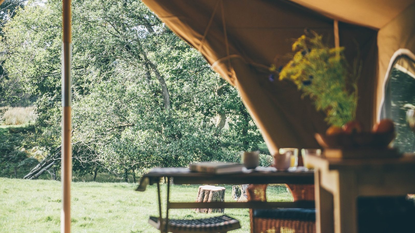 A cozy outdoor tent setup with a table and chairs is shown in the foreground. The scene looks out onto a green, grassy area with trees in the background. A vase of flowers and a bowl of fruit sit on a small table inside the Nantseren tent.