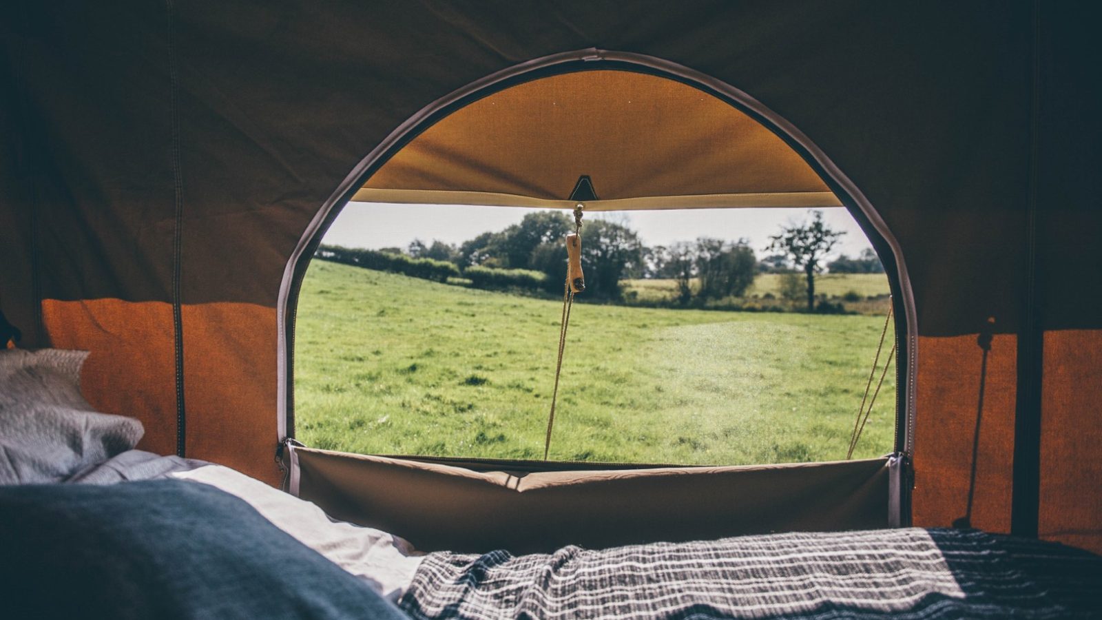 View from inside a Nantseren tent showing the open entryway looking out to a grassy field with trees in the distance. The inside of the tent is partially visible, including some bedding and blankets. The sky is clear and bright.