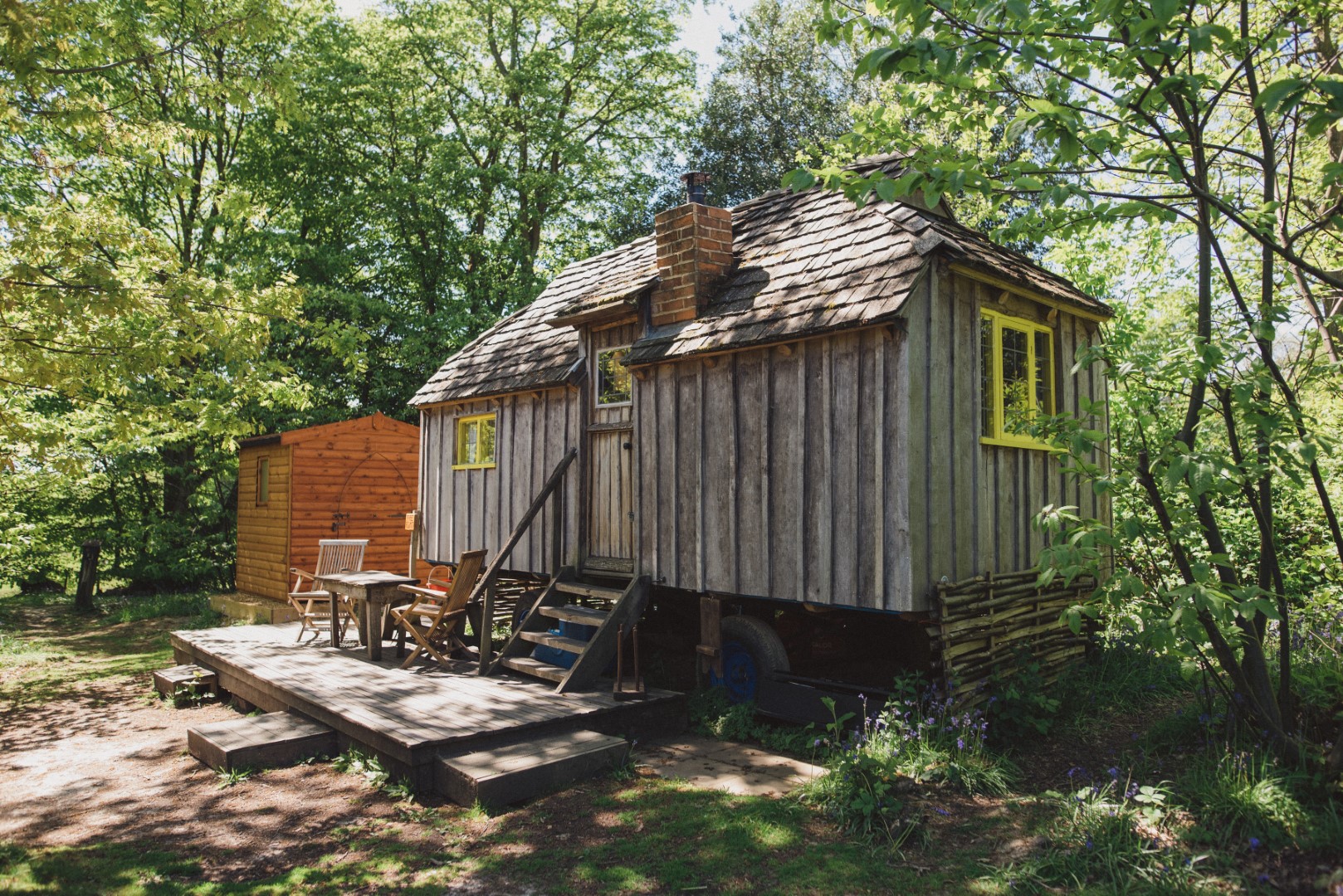 A small, rustic wooden cabin with a stone chimney and yellow window trim stands elevated on a forested lot. It features a front porch with wooden steps, outdoor chairs, and a table. Just 2 hours from London, this is one of the perfect places to stay to get away from the city. Another small wooden structure is visible in the background amidst lush greenery.