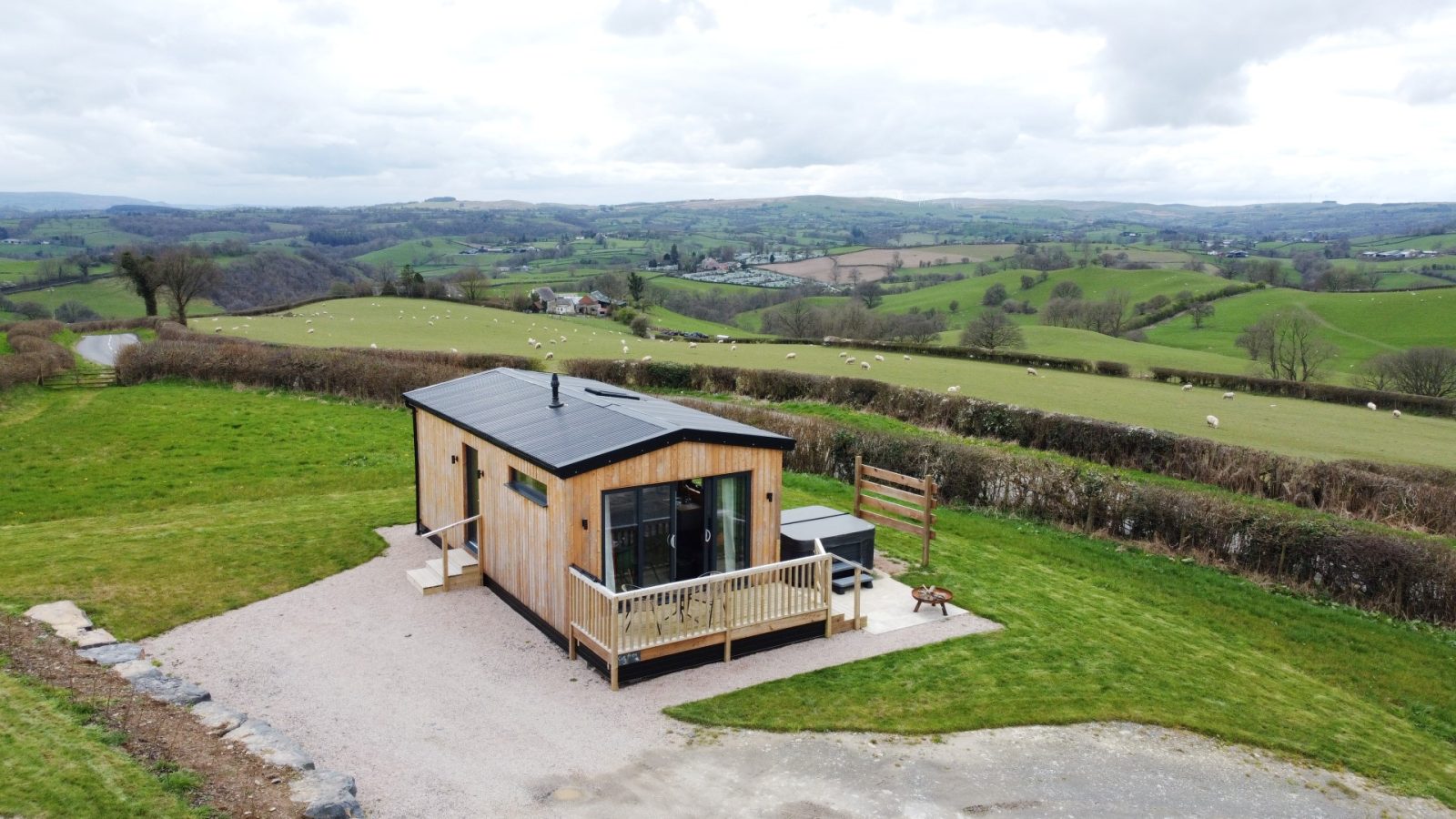 A small wooden cabin, part of the Belan View Lodges, features large windows that capture the essence of a grassy rural landscape with rolling hills in the background.