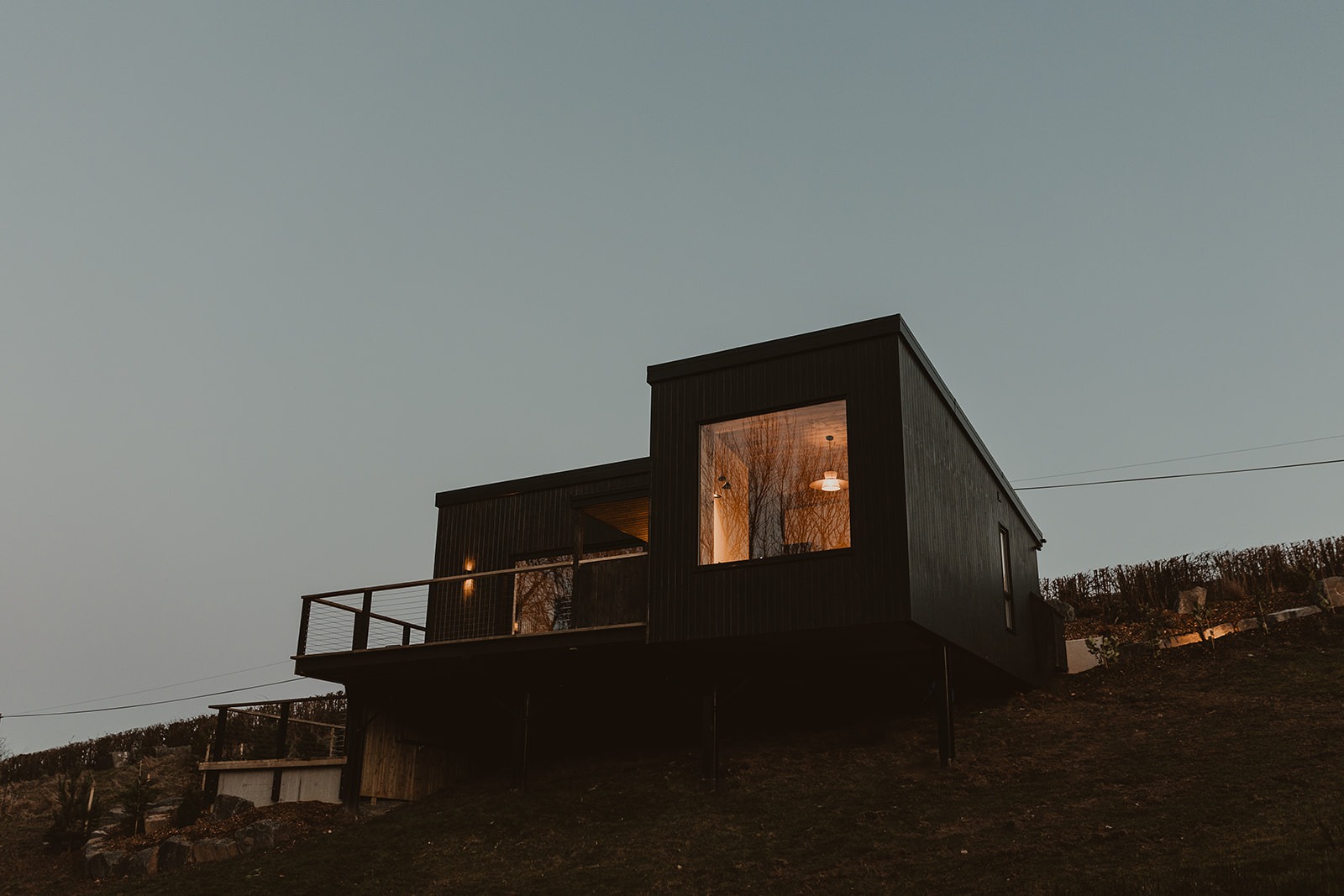 Modern hillside house with large windows, wooden cladding, and a terrace railing set against an evening sky, offering the intimate charm of cosy cabins for two.