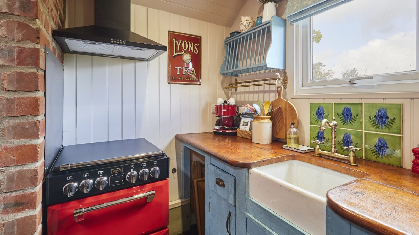 A compact kitchen with a red stove, blue cabinets, wooden countertops, a farmhouse sink under floral-tiled windows. Y Golchdy is the perfect escape for a romantic break for 2 in Wales