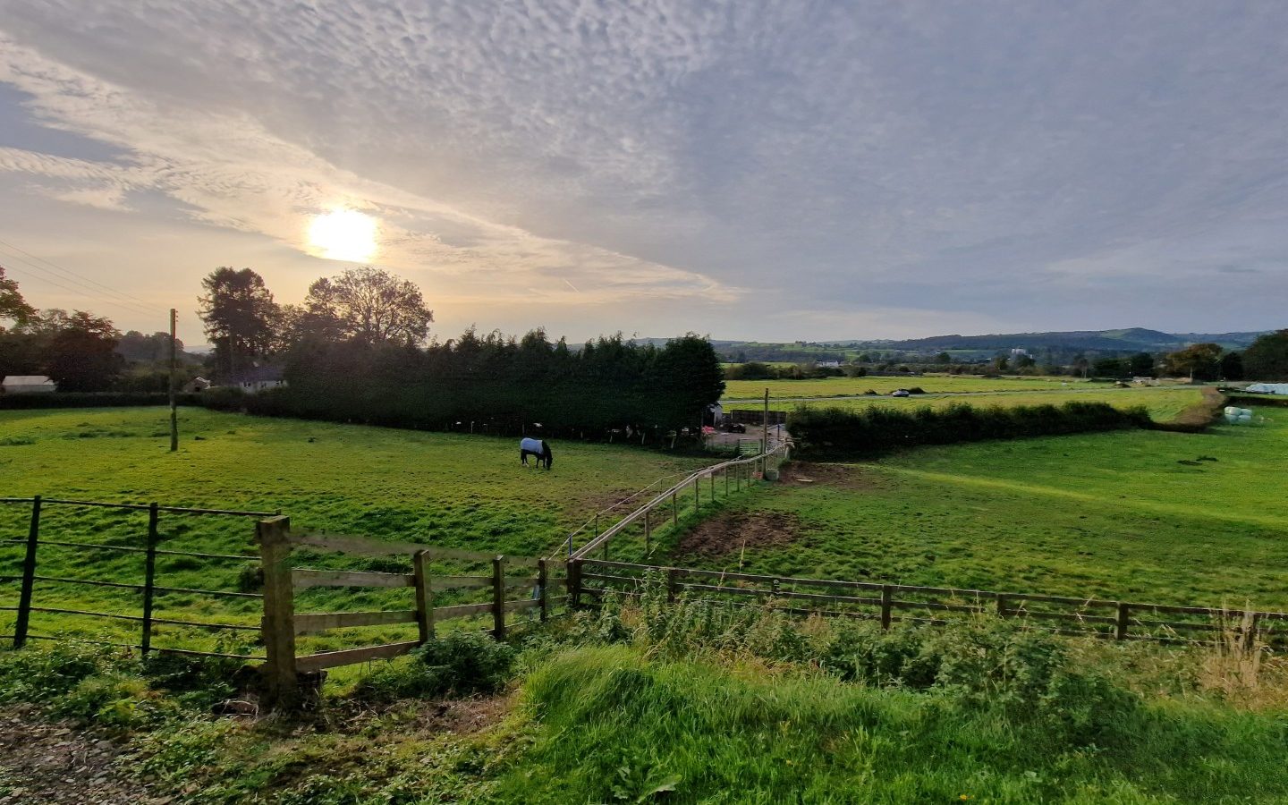 Pastoral landscape with green fields surrounding Y Golchdy, trees, and hills under a cloudy sky with the sun peeking through.