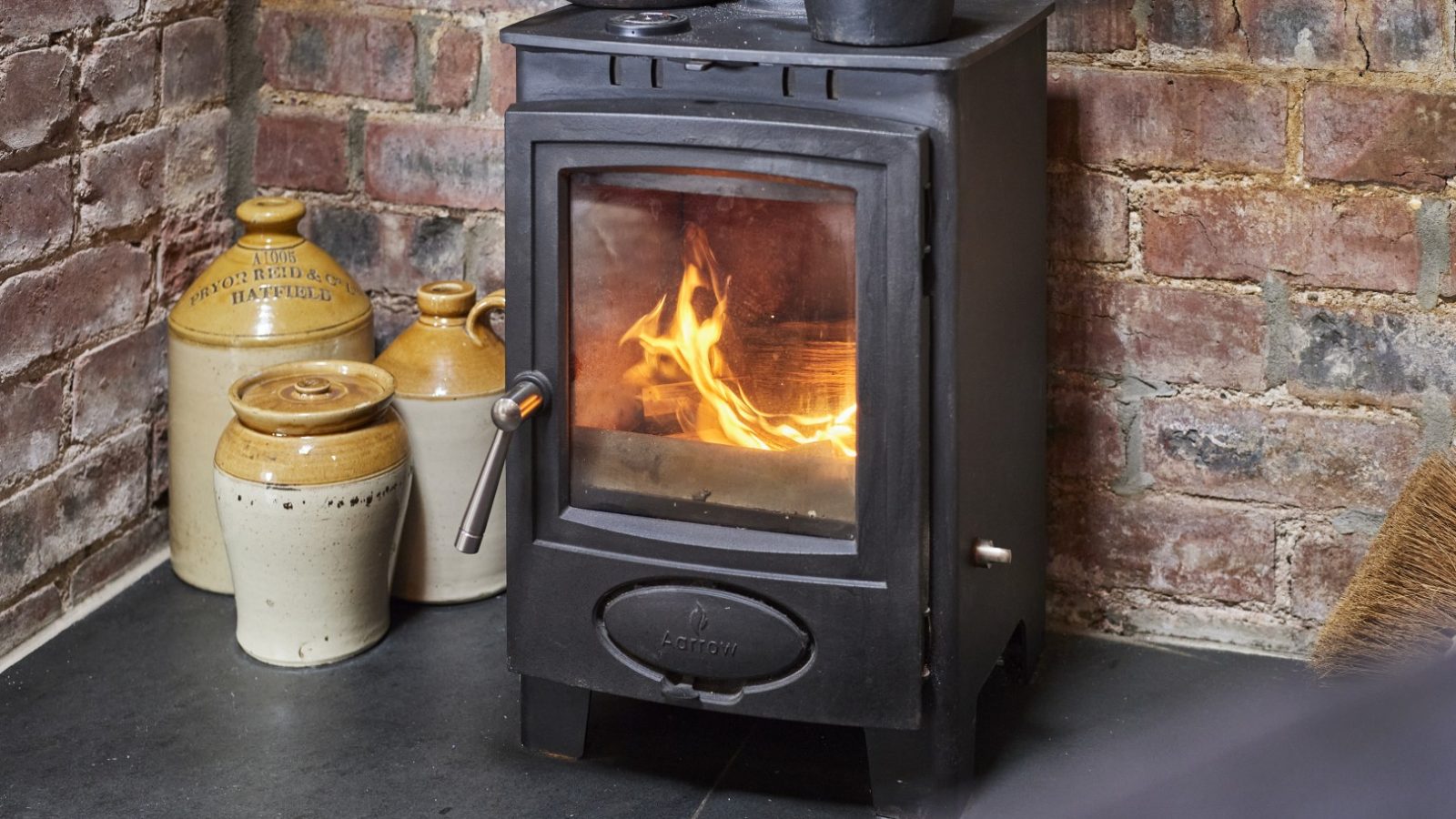 A small black stove with visible flames, set against a brick wall, with ceramic jugs nearby on the floor. Y Golchdy - a cosy cottage break for two in Wales