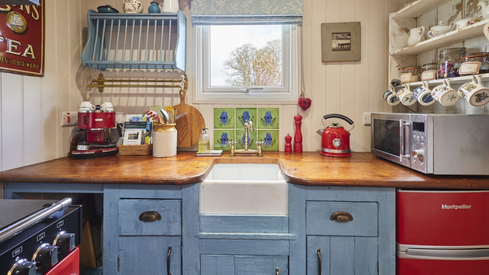 Cosy kitchen with blue wooden cabinets, red appliances, a farmhouse sink, and decorative items around a window. A romantic cottage break for 2 in Wales