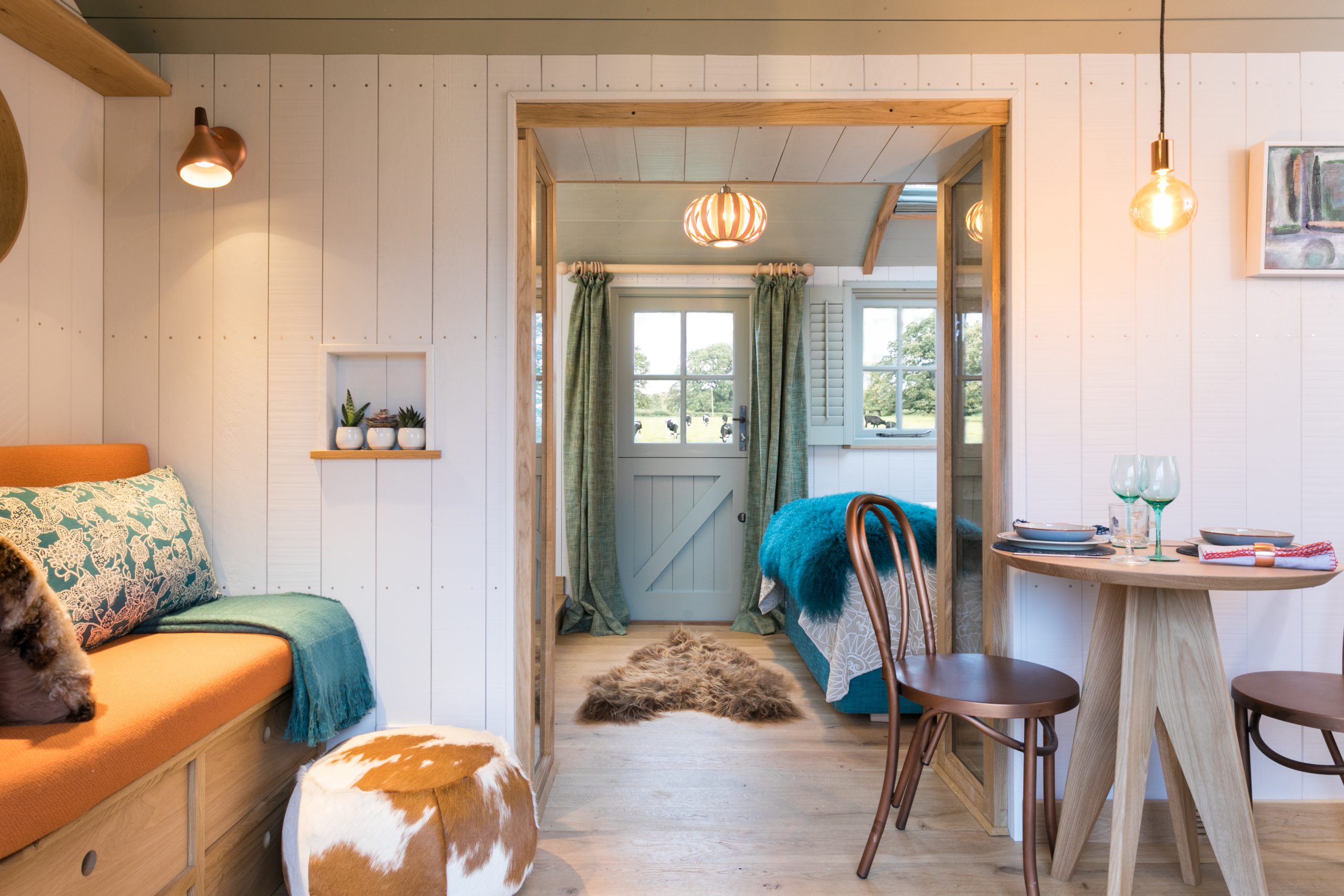 A cozy, well-lit tiny house interior with white paneled walls and wooden accents. In the foreground is a small dining area with two chairs and a round table. To the left, an orange built-in bench with cushions and a pouf adds value to the space. Beyond, a doorway reveals a bedroom. Perfect for maximizing holiday let profits.