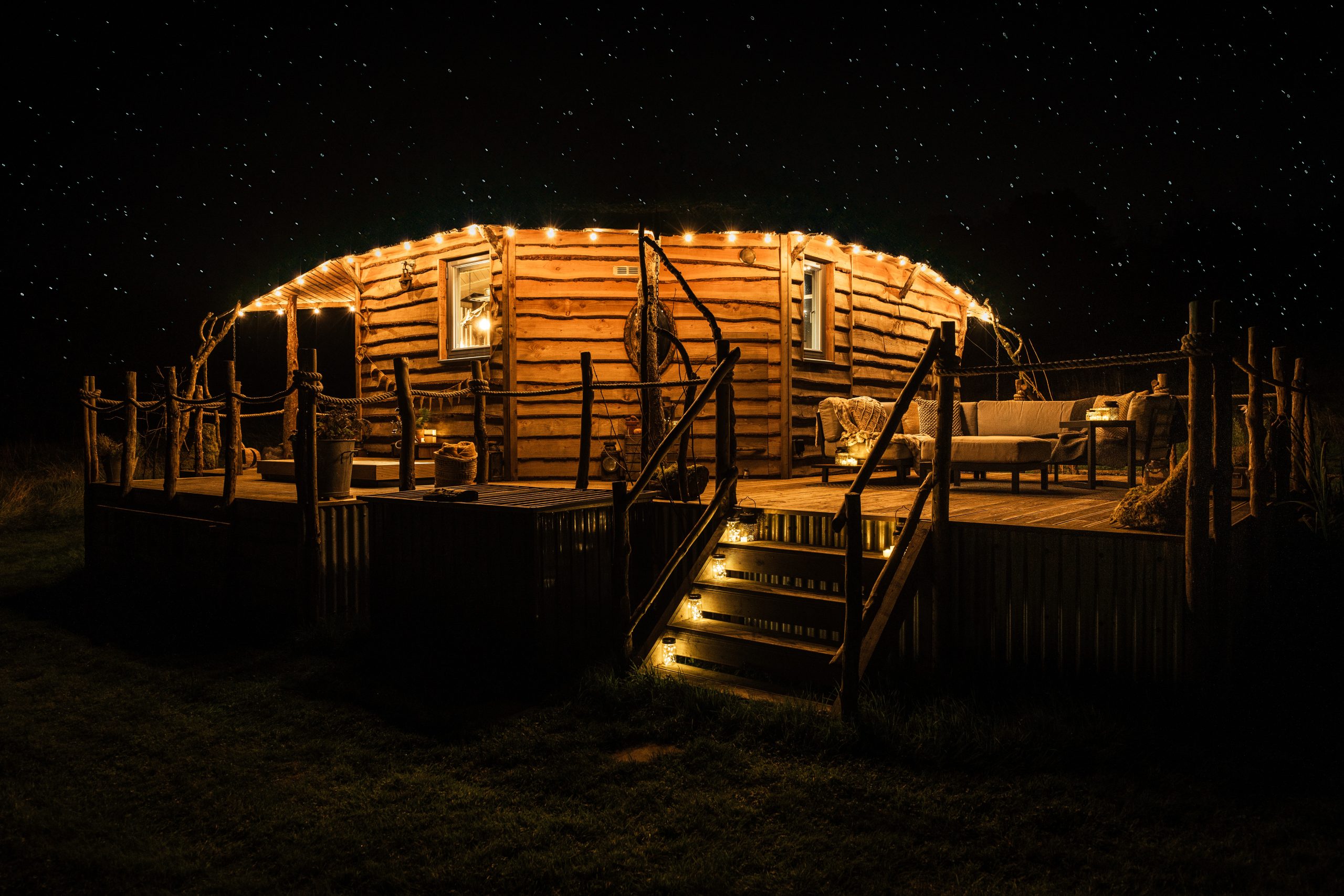 A cosy cabin for two, where a round wooden structure is gently illuminated by string lights on the porch, nestled under a starry night sky.